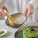 Crop anonymous cook mixing water and green food coloring using whisk on desk with stack of pancakes with blueberries and mint