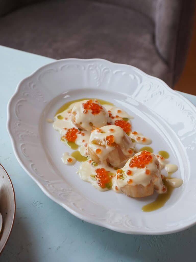 Close-up of a succulent scallop dish with creamy sauce and red caviar on an elegant ceramic plate.
